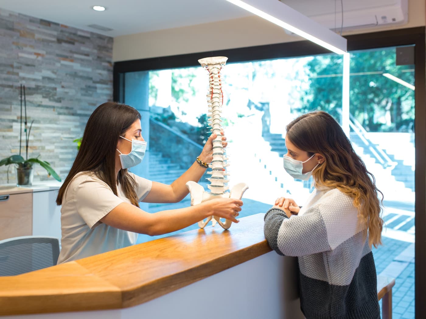 Recepcionista de clínica mostrando um modelo de coluna vertebral para uma paciente no balcão